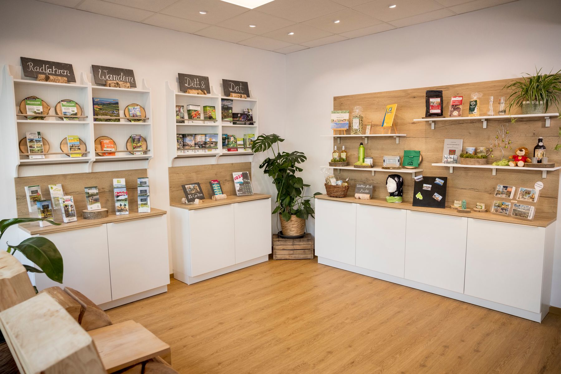 Ein modernes Besucherzentrum mit weißen Schränken, Holzregalen mit Broschüren, Karten und lokalen Produkten, Topfpflanzen und einem Holzboden mit heller Beleuchtung.